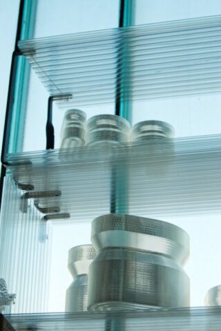 Transparente Regale aus Glas oder Acryl mit mehreren zylindrischen, durchsichtigen Behältern sind vor einem Fenster mit vertikalen, blau getönten Balken gestapelt.