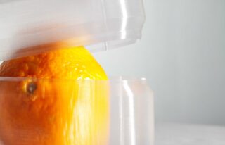 Eine Nahaufnahme einer Orange, die teilweise in einem durchsichtigen, starren Plastikbehälter mit strukturierter Oberfläche eingeschlossen ist.