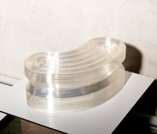 Ein durchscheinendes, nierenförmiges Acrylobjekt mit geschichteten Kurven sitzt auf einer reflektierenden weißen Oberfläche vor einer leicht strukturierten Wand.