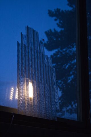 In der Abenddämmerung steht ein gemustertes Metallblech aufrecht vor einem Fenster, draußen leuchtet ein Licht und die Äste der Bäume sind zu sehen.