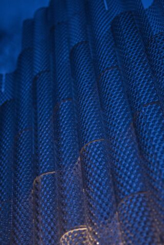 Nahaufnahme einer strukturierten, metallischen Oberfläche mit einem sich wiederholenden Muster, beleuchtet von blauem Licht, wodurch ein Gefühl von Tiefe und Abstraktion entsteht.