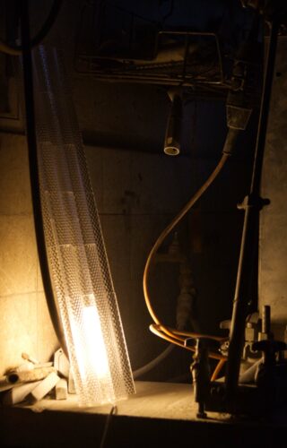 Eine Werkbank mit Werkzeugen, einer durch ein Drahtgeflecht geschützten Leuchtstoffröhre, einem aufgerollten gelben Schlauch und einer Metallklammer in einer schwach beleuchteten Werkstatt.