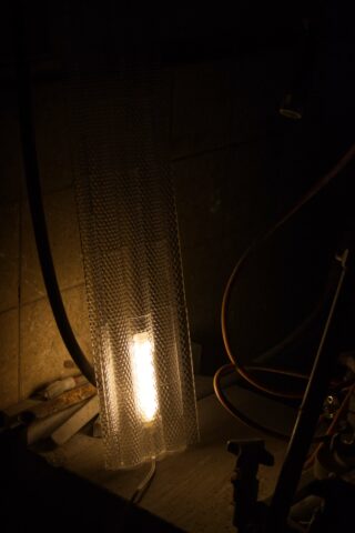 Eine Leuchtstoffröhre leuchtet hinter einem Maschendrahtgitter in einem dunklen, industriell anmutenden Raum, in dessen Nähe Kabel und Rohre sichtbar sind.