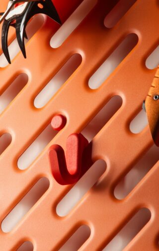Eine orangefarbene Lochplatte mit länglichen ovalen Löchern, an deren Rändern ein roter Haken und Teile von zwei Kletterwerkzeugen befestigt sind.