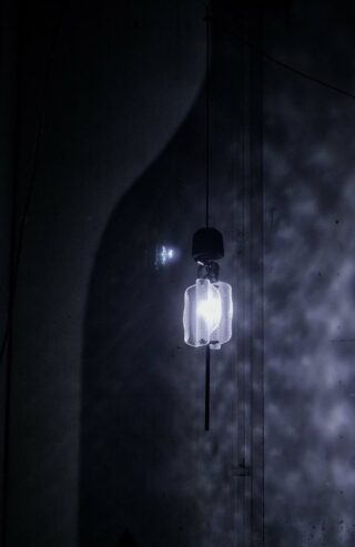 Eine einzelne beleuchtete Pendelleuchte hängt in einem dunklen Raum und wirft weiche Schatten und Muster auf die nahe Wand.