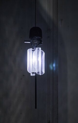 Eine moderne Pendelleuchte mit zylindrischem Netzschirm strahlt in einem schwach beleuchteten Raum ein kühles weißes Licht aus.