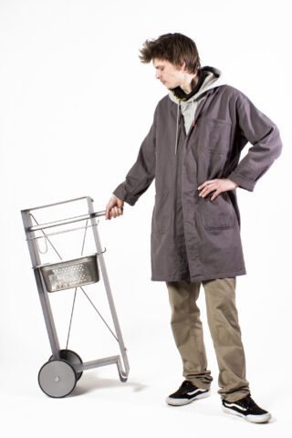 Eine Person in einem grauen Mantel und Khakihosen steht auf einem weißen Hintergrund und hält den Griff eines Metallwagens mit zwei Rädern und einem Korb.