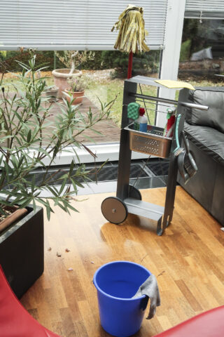 Ein Reinigungswagen mit Zubehör steht auf einem Holzboden neben einem blauen Eimer und einem Mopp in einem Raum mit großen Fenstern und Zimmerpflanzen.