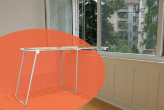 Ein zusammenklappbarer Wäscheständer mit weißen Metallbeinen steht in der Nähe großer Fenster in einem hellen Raum. Eine orangefarbene ovale Grafik überlagert einen Teil des Bildes.