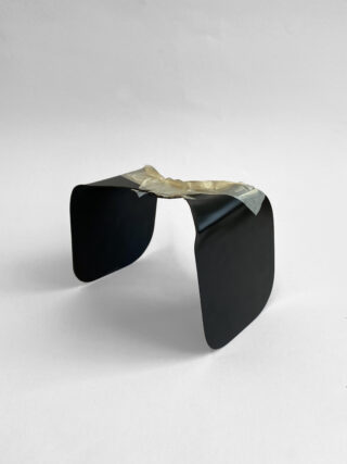 Auf einem schlichten weißen Hintergrund befindet sich ein kleines, gebogenes schwarzes Objekt mit quadratischen Kanten, das oben mit Klebeband zusammengehalten wird.