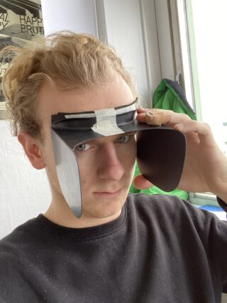 Eine Person trägt ein selbstgemachtes Visier aus schwarzem Plastik und Klebeband, hält es an ihrer Stirn und hat einen Verband am Finger.