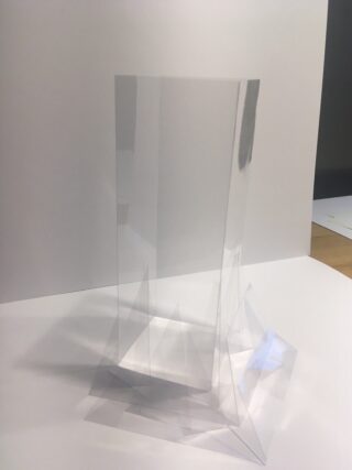 Eine hohe, transparente geometrische Struktur aus durchsichtigen Kunststoffplatten steht auf einer weißen Fläche vor einem weißen Hintergrund.