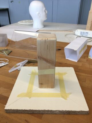 Ein rechteckiger Holzblock ist mit Klebeband aufrecht auf einem quadratischen Brett befestigt, umgeben von Kunstmaterialien auf einem Arbeitstisch. Im Hintergrund ist ein weißes Kopfmodell zu sehen.