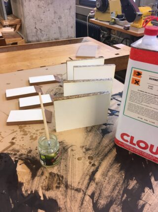 Auf einer Werkbank in einer Holzwerkstatt sind mehrere Spanplatten, ein Leimpinsel in einem kleinen Gefäß und ein großer Behälter mit Klebstoff angeordnet.