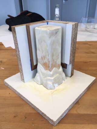 Eine selbstgemachte Form umgibt ein zylindrisches Objekt mit strukturierter Basis, das auf einem quadratischen Brett auf einer hölzernen Arbeitsfläche steht.