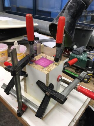 Eine hölzerne Kastenform wird von roten und schwarzen Klammern auf einer Werkbank zusammengehalten, auf der überall Werkzeuge und Materialien verstreut herumliegen.