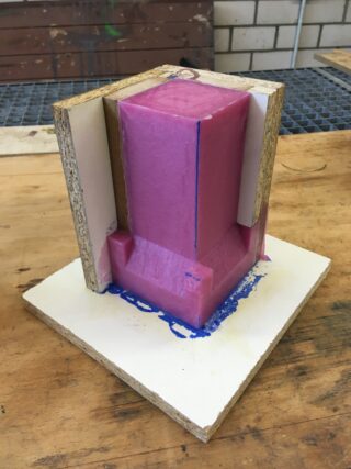 Eine rosafarbene rechteckige Form mit scharfen Kanten ist von Holzbrettern umschlossen und steht auf einem weißen Sockel auf einer Werkbank. An der Unterseite ist blaues Material herausgesickert.