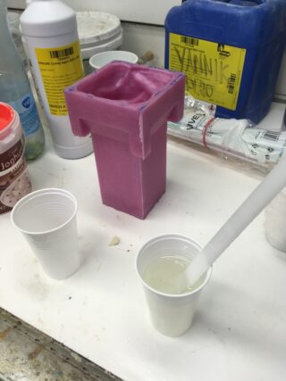 Neben einer rosa Silikonform und anderen Behältern auf einer Arbeitsfläche wird in einem Plastikbecher mit einem Rührstab eine klare Flüssigkeit gemischt.