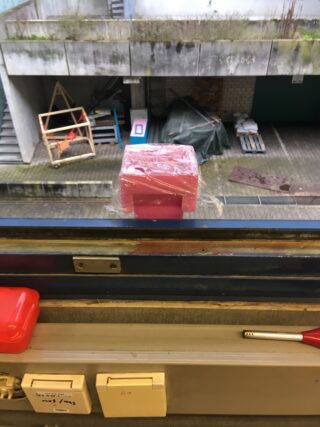 Auf einem Fensterbrett steht ein rosa, rechteckiger, in Plastik eingewickelter Gegenstand. Draußen sind in einem Betonhof Treppen, weggeworfene Bretter und Schutt zu sehen.