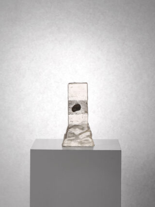 Eine klare Glasskulptur mit rauer Textur und einem darin eingebetteten kleinen dunklen Objekt, ausgestellt auf einem weißen Sockel vor einem schlichten grauen Hintergrund.