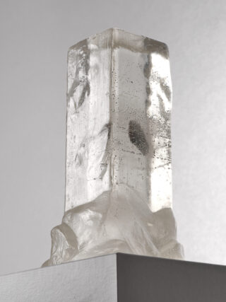 Eine klare, rechteckige Kristallskulptur mit rauen, strukturierten Oberflächen wird auf einem weißen Sockel vor einem schlichten grauen Hintergrund präsentiert.