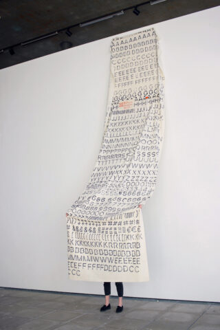 Eine Person steht hinter einem großen, vertikal hängenden Stoff, der mit handgeschriebenen Buchstaben bedeckt ist, hauptsächlich den Großbuchstaben C, D, E und G, die vor einer weißen Wand in einer Galerie ausgestellt sind.