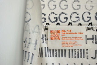 Nahaufnahme eines Textilkunstwerks mit großen schwarzen Buchstaben und einem orangefarbenen Etikett mit Angaben zum Titel des Stücks, den Mitwirkenden, den Abmessungen und einem QR-Code.