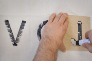 Eine Person verwendet eine Schablone und einen Marker, um den Buchstaben „J“ auf Stoff zu zeichnen, neben die bereits mit der Schablone aufgemalten Buchstaben „V“ und „O“.