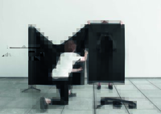 Eine Person kniet und schneidet schwarzen Stoff von einer Rolle, während eine andere Person ein großes ausgeschnittenes Stück mit Rucksackgurten hochhält. Beides liegt auf einem gefliesten Boden vor einer schlichten weißen Wand.