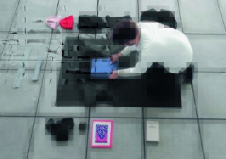 Eine Person kniet auf einer Matte und ordnet einen blauen Ordner, umgeben von verschiedenen Gegenständen, darunter Taschen, Hüte, zwei Bücher und kleine persönliche Gegenstände auf Betonfliesen.