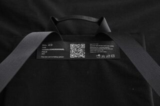 Ein schwarzer Stoffartikel mit zwei Riemen verfügt über ein weißes Etikett mit Pflegehinweisen, Materialdetails und einem QR-Code für Faltmöglichkeiten.