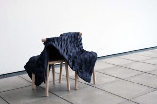 Auf einem gefliesten Boden steht ein Holzstuhl, dessen Rückenlehne und Sitzfläche mit einem dunklen, gewebten Stoff bedeckt sind.