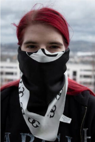 Eine Person mit langen roten Haaren trägt ein schwarz-weißes Bandana, das Nase und Mund bedeckt, und steht im Freien mit Gebäuden und einem bewölkten Himmel im Hintergrund.