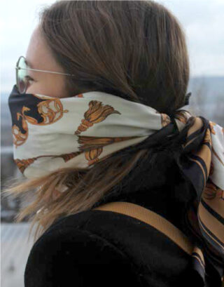 Person mit Sonnenbrille und einem gemusterten Schal, der um ihr Gesicht gebunden ist und Nase und Mund bedeckt, vor einem unscharfen Außenhintergrund.