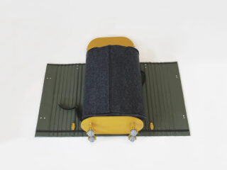 Ein gelb-graues zylindrisches Objekt mit Metallbeschlägen liegt auf einer grünen, gerippten Matte mit Löchern vor einem schlichten weißen Hintergrund.