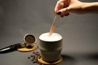 Eine Hand mit dunklem Nagellack hält eine Pipette über einem schaumigen Getränk in einer Keramiktasse, daneben stehen gemahlener Kaffee, Kaffeebohnen und ein Siebträger auf Untersetzern.