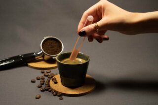 Eine Hand taucht eine Pipette in eine Tasse Kaffee auf einem Korkuntersetzer, daneben befinden sich Kaffeebohnen und gemahlener Kaffee in einem Siebträger.