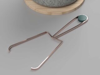 Eine Küchenzange aus Metall mit einem blaugrünen, runden Griff liegt auf einer grauen Oberfläche neben einem runden Holzbrett, auf dem eine Steinschüssel steht.