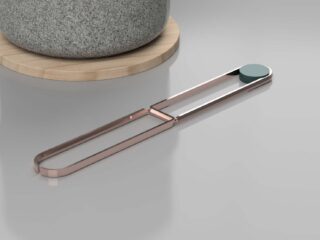 Ein metallisches Küchenwerkzeug mit einer rechteckigen Schlaufe und einem runden Pad liegt auf einer grauen Oberfläche neben einem Steintopf auf einem Holzuntersetzer.