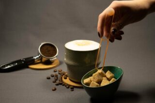 Eine Hand hält eine Zuckerzange über eine Schale mit Zuckerwürfeln, daneben stehen auf einer grauen Fläche eine Tasse Cappuccino und ein Siebträger mit gemahlenem Kaffee.
