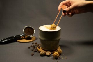 Eine Hand hält Essstäbchen, um einen Trüffel in ein schaumiges Getränk neben Kaffeebohnen, gemahlenem Kaffee und Gewürzen auf einem grauen Hintergrund zu tauchen.