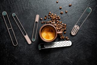 Eine Tasse Espresso steht auf einer dunklen Oberfläche, umgeben von verstreuten Kaffeebohnen und mehreren Kaffeeutensilien aus Metall.