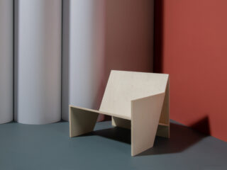 Ein minimalistischer Holzstuhl mit abgewinkelten Paneelen steht auf einem blaugrauen Boden vor dem Hintergrund weißer Zylinder und einer rötlichen Wand.