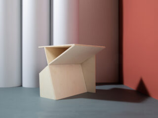 Ein minimalistischer, geometrischer Holzstuhl mit eckigen Linien steht auf einem grauen Boden vor einem Hintergrund aus weißen zylindrischen und roten flachen Paneelen.