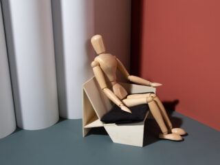 Eine hölzerne Künstlerpuppe sitzt auf einem modernen, eckigen Stuhl mit einem schwarzen Kissen vor einem Hintergrund aus weißen Zylindern und einer roten Wand.