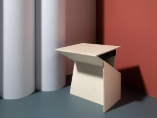 Ein minimalistischer, eckiger beiger Hocker steht auf einem grauen Boden neben zylindrischen weißen Säulen und einer roten Wand.