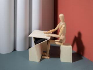 Eine hölzerne Künstlerpuppe sitzt an einem geometrischen Schreibtisch mit schräger Oberfläche vor einer roten Wand und zylindrischen weißen Objekten.