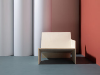 Ein minimalistischer Holzstuhl steht auf einem grauen Boden mit zylindrischen weißen Säulen und einer gedämpften roten Wand im Hintergrund.