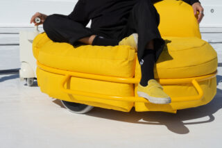 Eine schwarz gekleidete Person sitzt auf einem leuchtend gelben, gepolsterten, runden Gartenstuhl mit Rollen auf einer weißen Fläche.