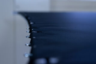 Nahaufnahme eines dunklen Stoffes, der mit Metallstiften am Rand gespannt und befestigt ist. In der Mitte ist ein Riss sichtbar. Der Hintergrund ist unscharf.